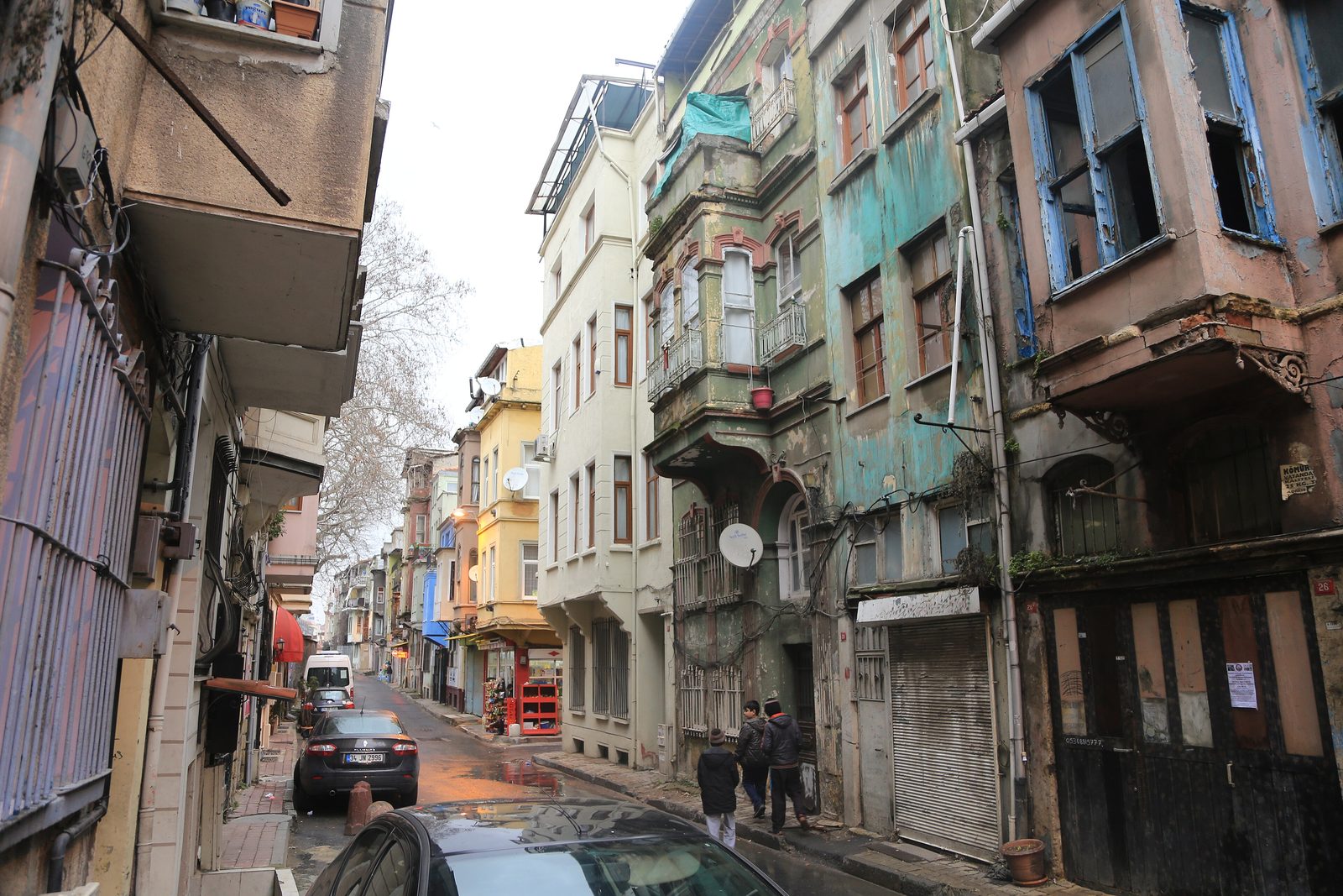 A narrow cobbled street in Balat lined with multi-storey timber houses in layered weathered colours — ochre yellow, faded teal-green, dusty pink, sage — with bay windows, wrought-iron balconies, and a glimpse of the Phanar slope rising at the end of the lane