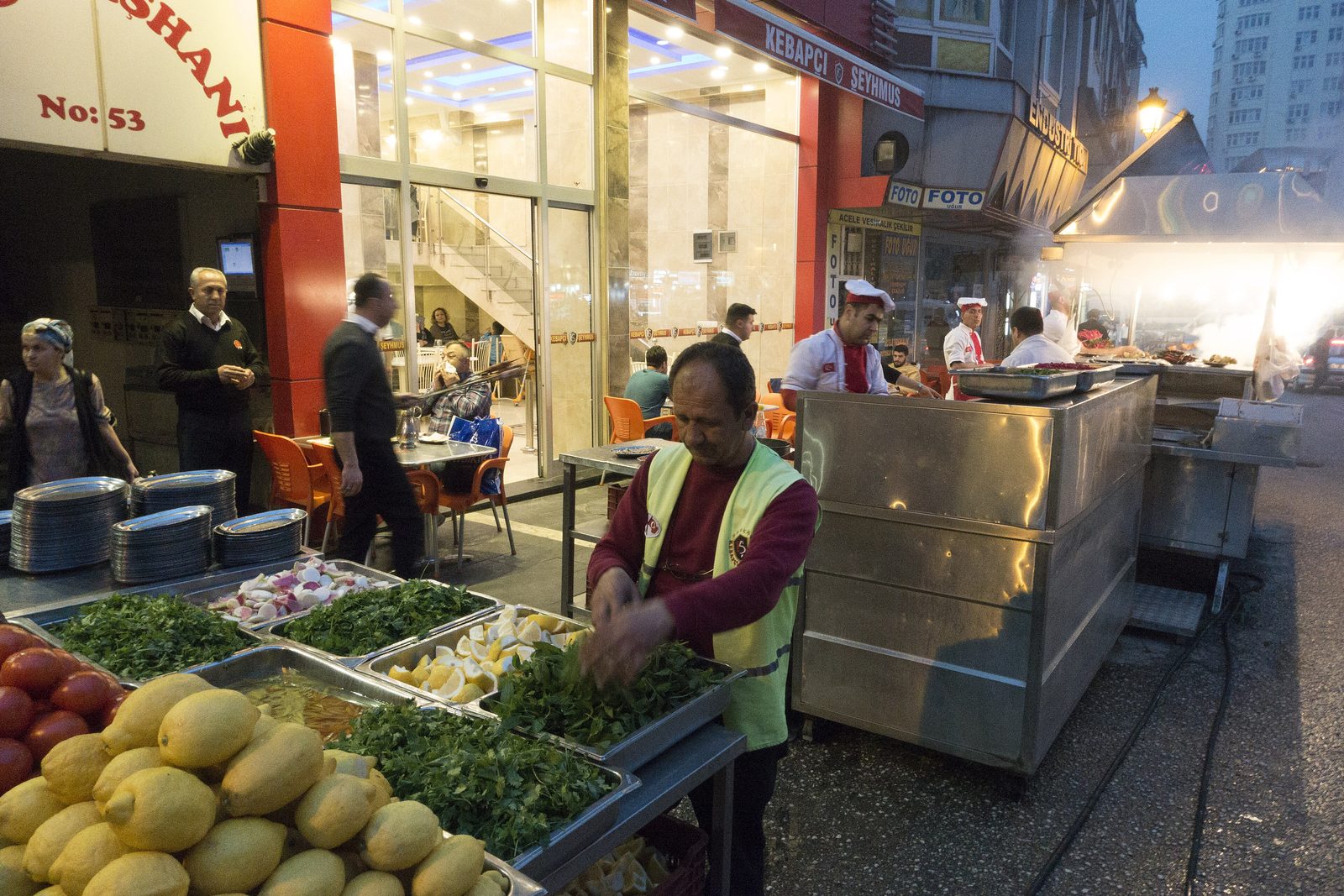 An Istanbul kebabçı at dusk — the working open-air grill station in front of the restaurant, a smoking charcoal grill on the right with chefs and a runner in white coats and red collars, foreground trays of trimmed parsley, lemons, sliced onions and pickled chillies on a stainless-steel prep table, a worker in a red jacket and high-vis vest tending the herbs