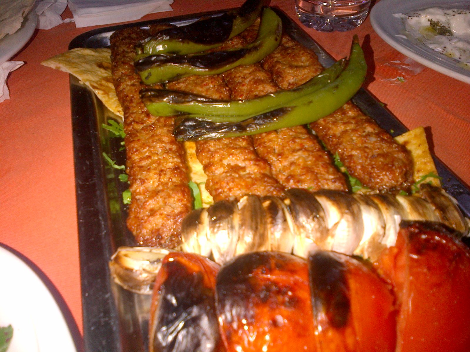Two long Adana kebabs on flat metal skewers laid on a black platter over warm lavaş bread, with the rough irregular hand-chopped texture of the meat clearly visible, alongside grilled long green peppers, charred tomatoes and a grilled long aubergine