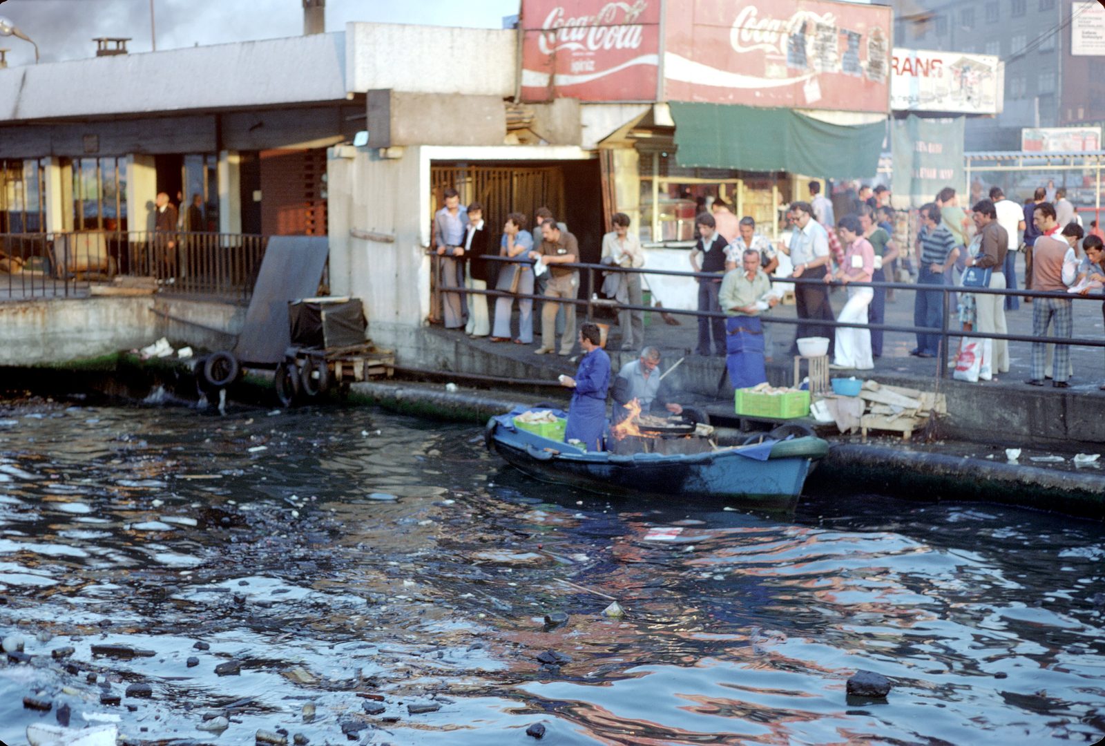 The ornately-decorated wooden balık-ekmek boats moored at the Eminönü waterfront on the Golden Horn — gold-painted hulls, charcoal grills running open-flame, vendors in red waistcoats and white shirts, with a queue of customers waiting at the rail and the Galata Bridge in the background