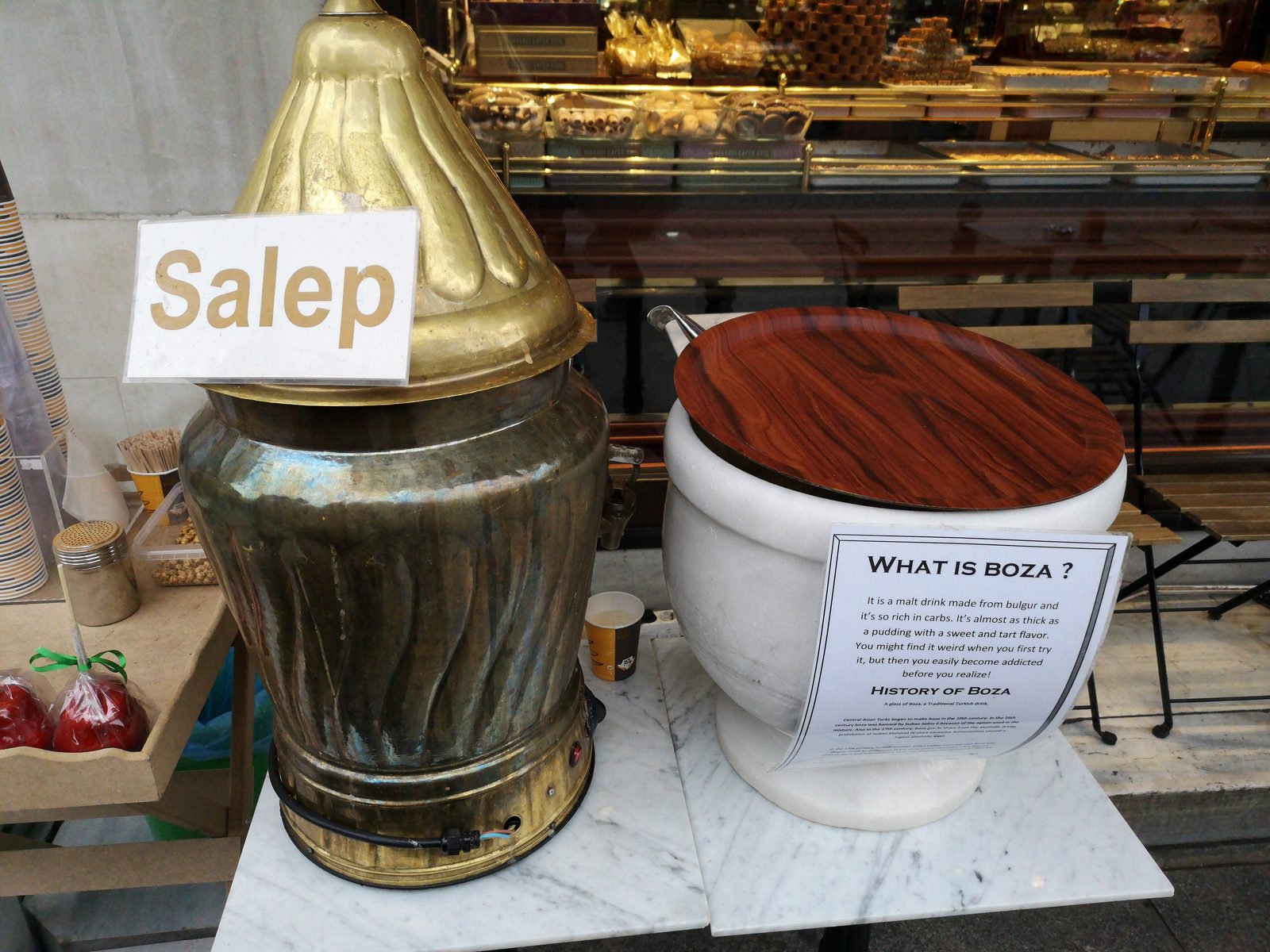 Two traditional Turkish drink urns on a marble surface in front of a shop window: on the left, a tall brass-and-copper urn with a pointed conical lid labeled Salep; on the right, a white ceramic urn with a wooden lid bearing a printed What is Boza notice