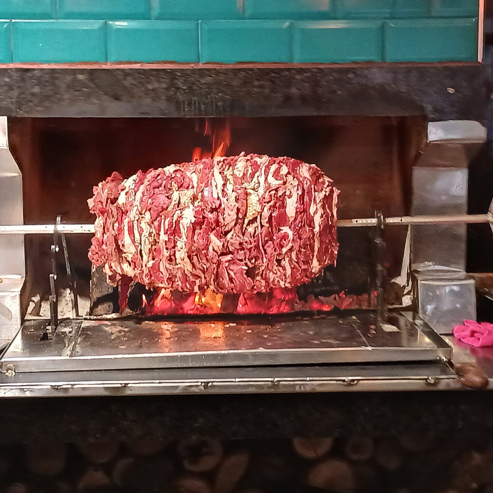 A horizontal cağ kebabı setup — long layers of marinated lamb stacked vertically and mounted on a horizontal rotating iron rod above glowing wood embers, with a green-tiled hearth surround behind