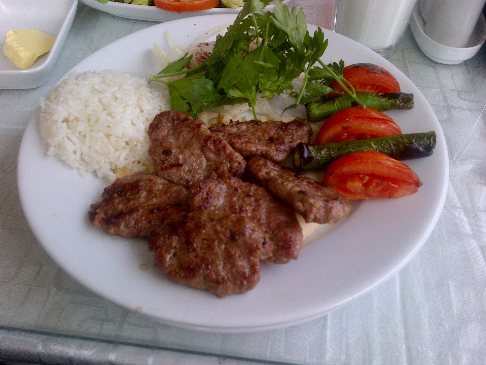 A plate of Turkish köfte — five short flat charcoal-grilled meatballs in the centre, a mound of white rice on the side, two charred long green peppers, three wedges of red tomato, fresh parsley, on a white round plate at a marble café table