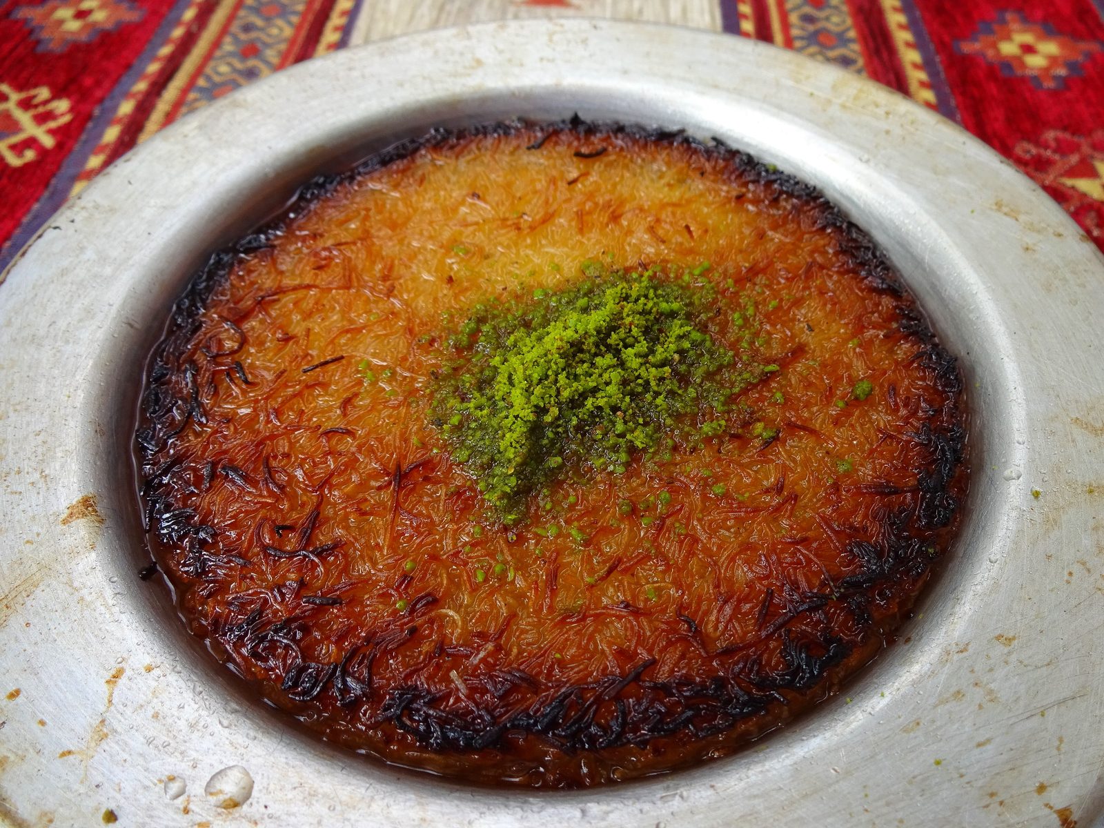 A small copper pan of freshly-baked künefe with the shredded-kataifi pastry baked golden and crisp on top, melted white cheese visible underneath at the broken edge, drizzled with sugar syrup, sprinkled with ground pistachio