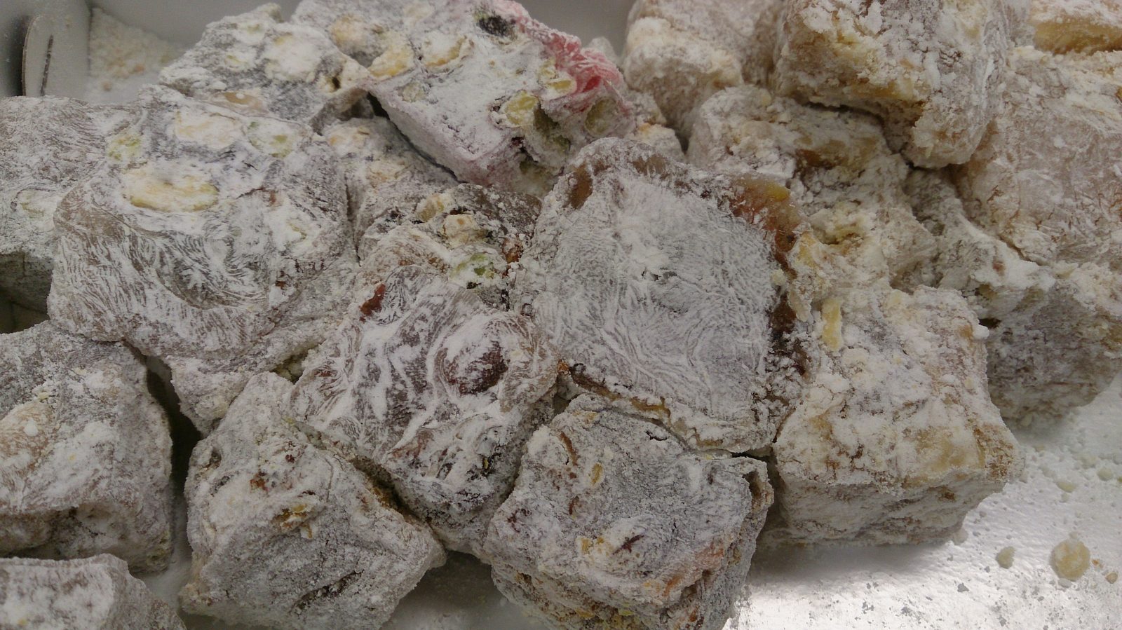 Hand-cut pieces of lokum piled together and heavily dusted in powdered sugar — the irregular cube shapes clearly visible, the natural muted tones of rose-amber, walnut-brown and mastic-white catching the light, with pistachio pieces embedded in some of the cubes