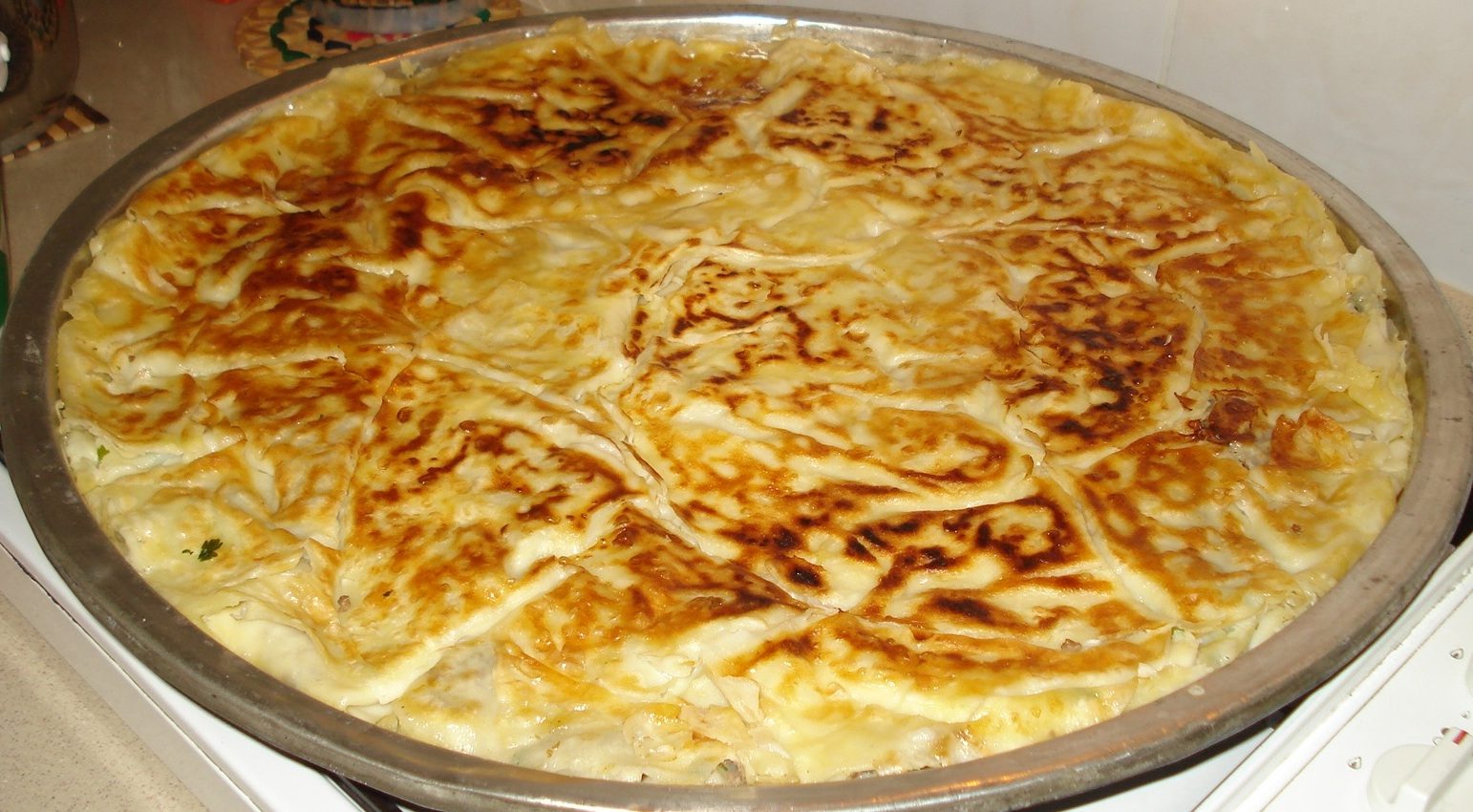 A whole round tray of freshly-baked su böreği on a metal pan — the golden-brown crinkled top layer is the defining surface, with the multi-layered cross-section visible at the cut edge of a wedge in the foreground