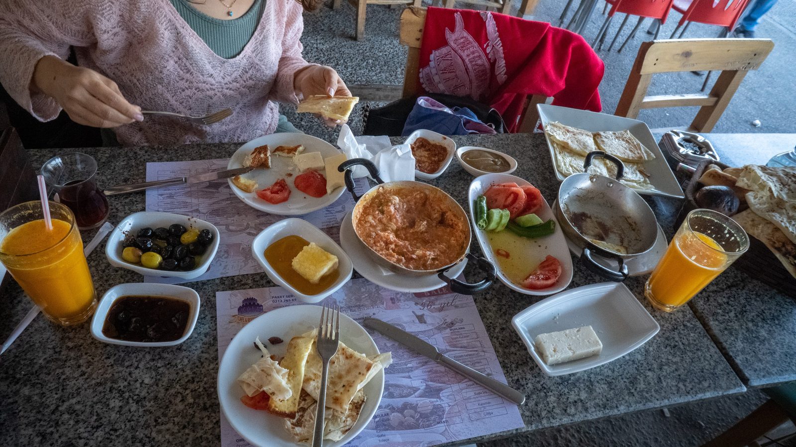An overhead view of a real Turkish kahvaltı spread on a wooden table — a copper pan of menemen, eggs, white cheese, sliced tomato and cucumber, olives, kaymak with honeycomb, jams, simit, bread and tulip glasses of black tea, with the Bosphorus visible through a window in the background