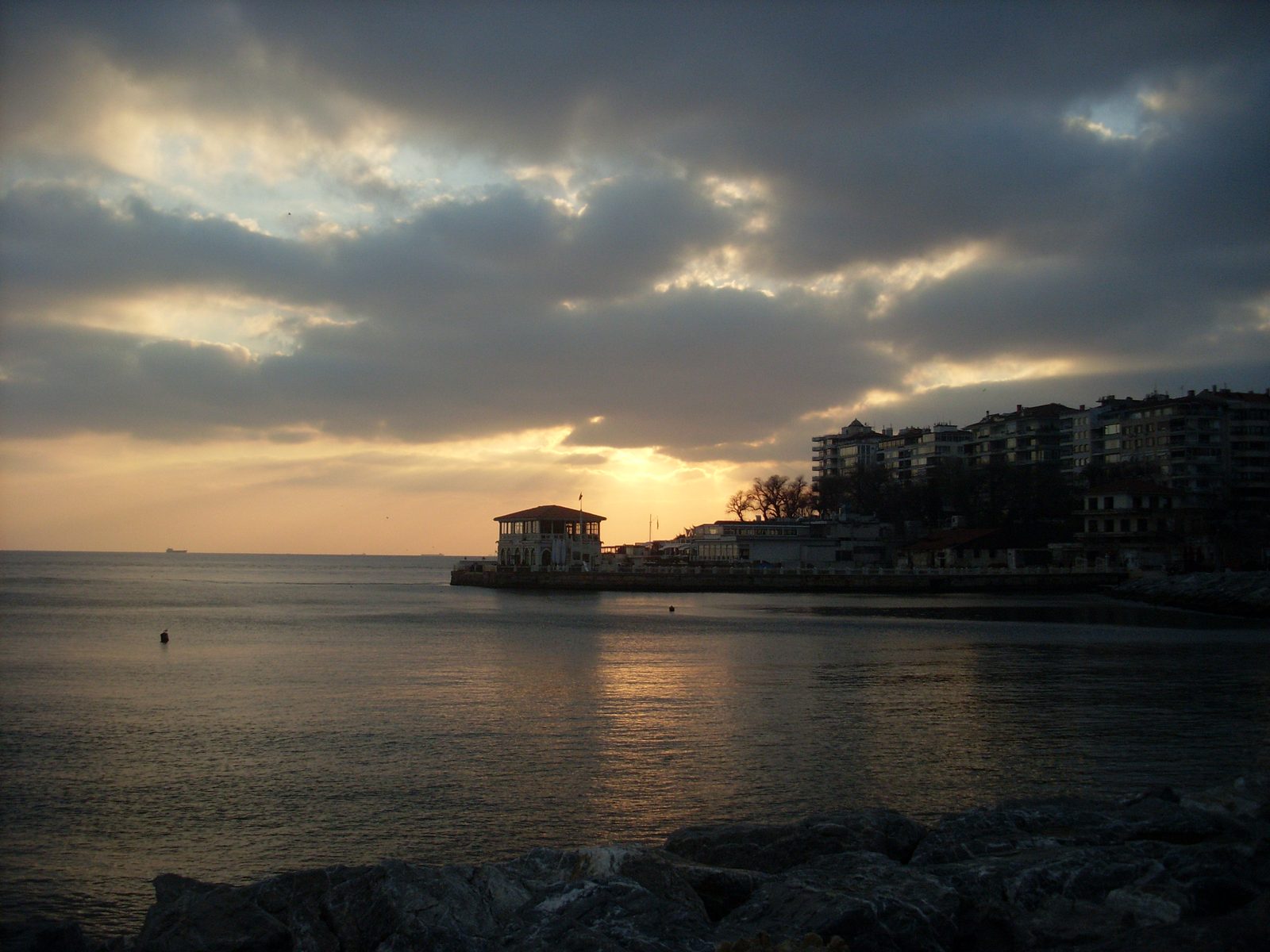 The Moda waterfront in Kadıköy at sunset, with the historic Moda iskele teahouse sitting on its small headland over the Sea of Marmara, mansion-blocks of the neighbourhood on the right, and the sun setting low over the strait