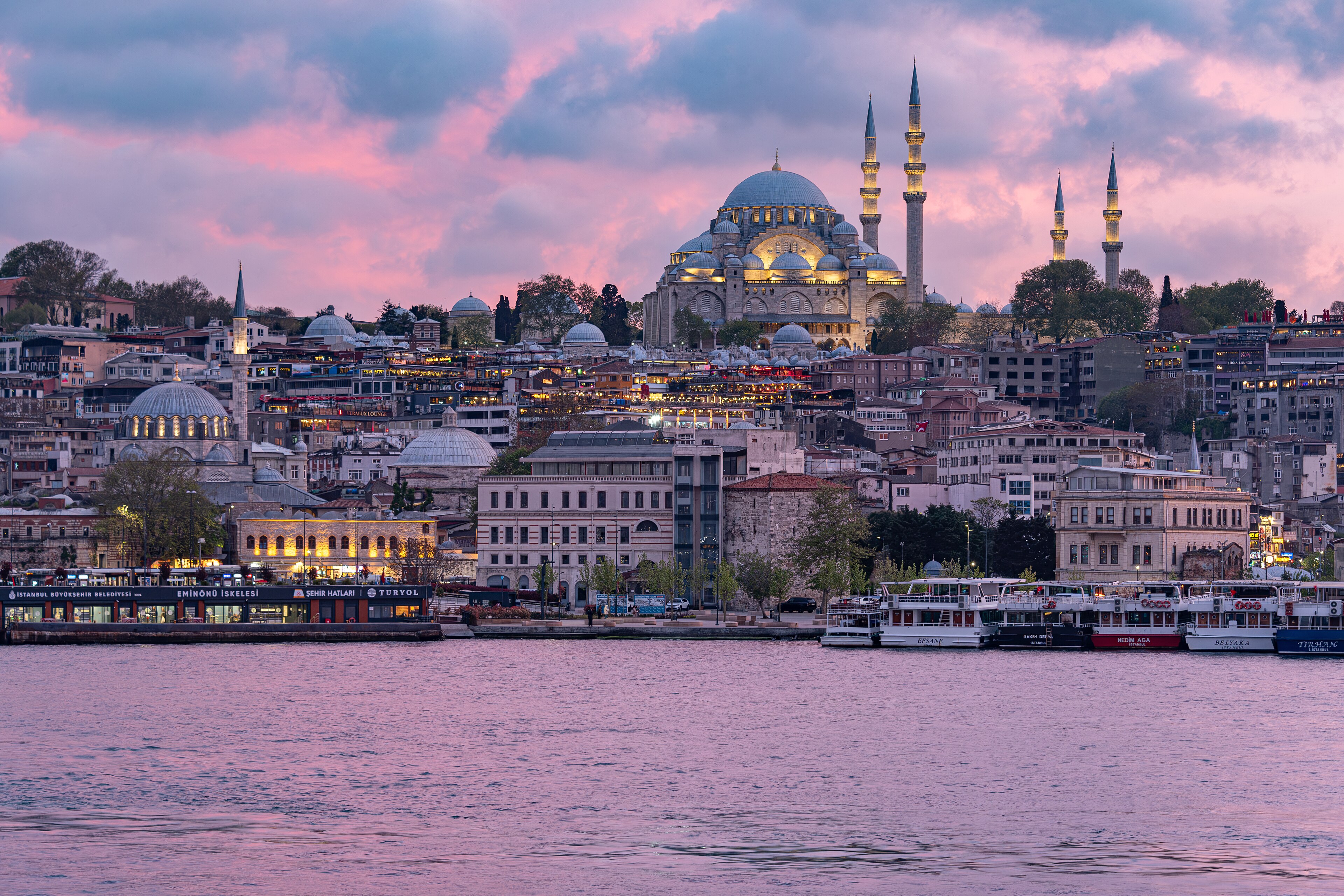 The view from Karaköy across the Golden Horn at sunset, with the Süleymaniye Mosque dominating the historic peninsula skyline and ferry boats moored at the Eminönü iskele