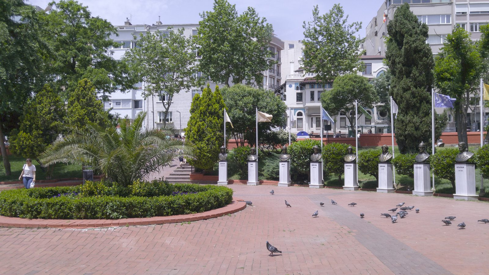 Nişantaşı Park on a bright afternoon — a small landscaped square with a row of bronze busts of Turkish historical figures on white marble plinths, plane trees and cypresses, brick paving, mansion-block façades behind, and pigeons walking the path