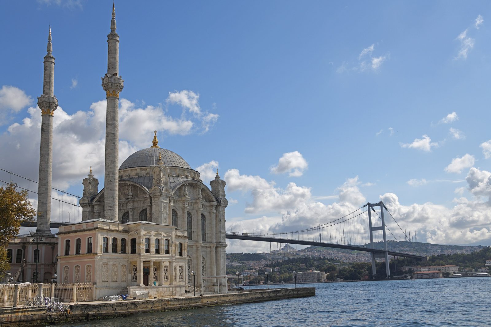 The Mecidiye Camii on the Ortaköy waterfront square at golden hour, its slender twin minarets and ornate Neo-Baroque dome sitting on a small headland directly on the Bosphorus, with the suspended deck of the 15 Temmuz Şehitler (1973 Bosphorus) Bridge arcing across the strait behind