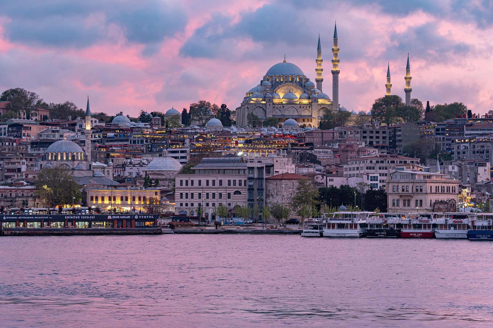 The Galata Bridge over the Golden Horn at sunset — the lower-deck fish-meyhane row visible underneath, the rod-fishermen on the pavement above, the silhouette of the historic peninsula's domes and minarets on the far bank