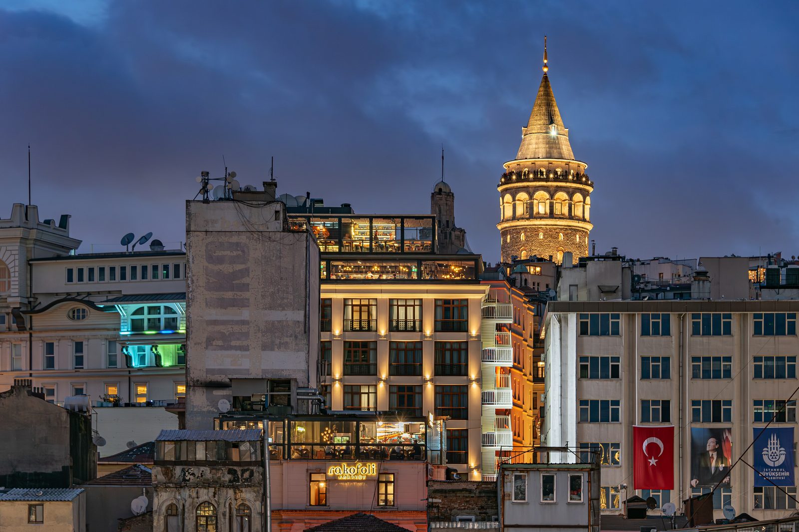 The Galata Tower at dusk above the rooftop terraces and lit dining rooms of the Beyoğlu/Karaköy district below, with the sky turning deep blue and Turkish flags hanging on the building facades