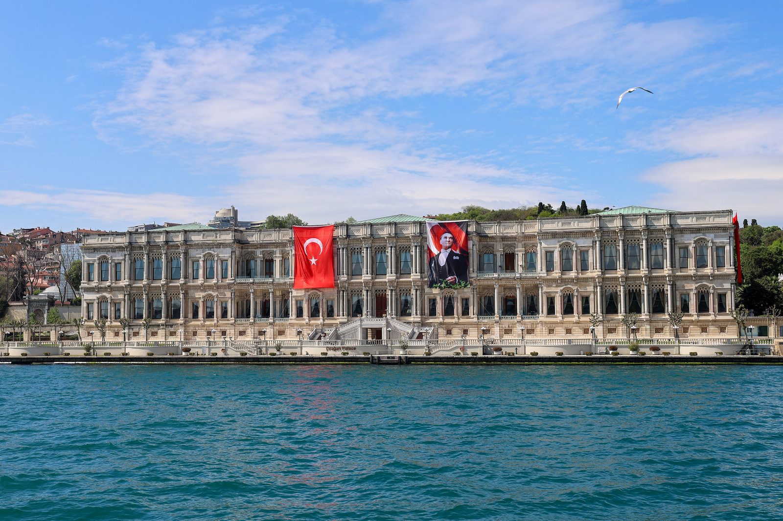 The Çırağan Palace seen from the Bosphorus — the long white marble Ottoman waterfront facade with arched windows, two large flags hanging over the central portico, and the strait in the foreground