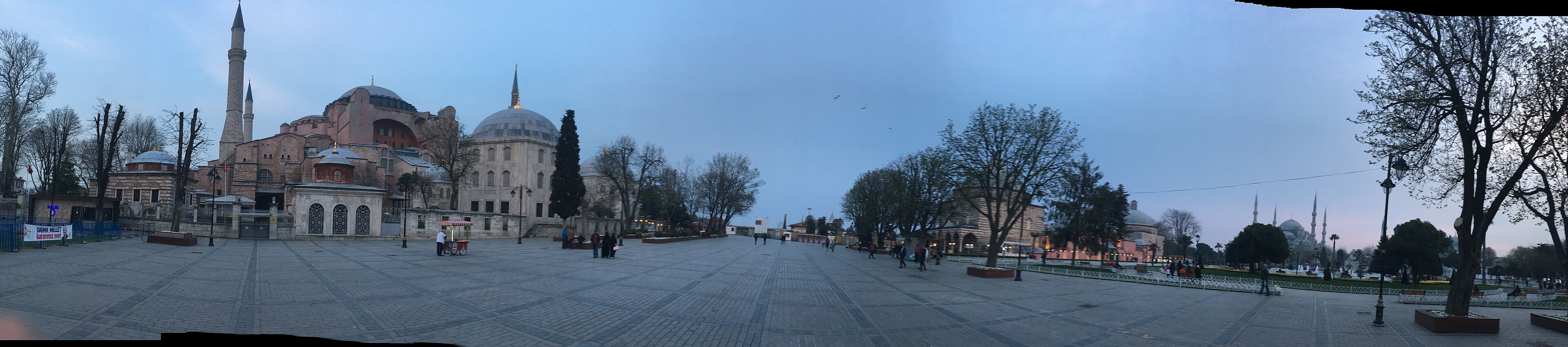 A wide-angle panorama of the Hippodrome of Constantinople (Sultanahmet Meydanı) — the long open public square between the Blue Mosque and the Hagia Sophia, with the Egyptian obelisk and the Serpent Column visible in the central axis and the surrounding old-city streets